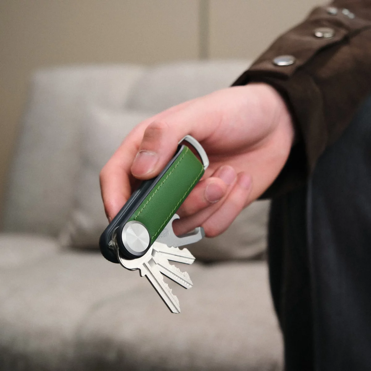 Hand hält den Orbitkey Key Organiser Tracker Leder in Waldgrün mit ausgeklappten Schlüsseln vor einem Sofa