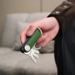 Hand hält den Orbitkey Key Organiser Tracker Leder in Waldgrün mit ausgeklappten Schlüsseln vor einem Sofa