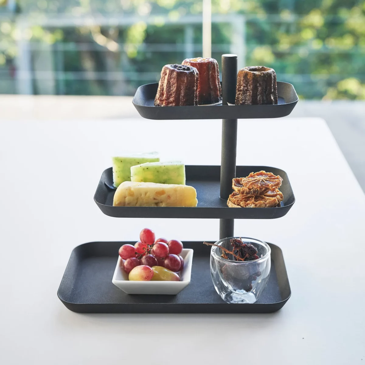 Yamazaki Etagere TOWER 3-stöckig drehbar in Schwarz, von vorne vor dem Fenster mit Früchten und Törtchen
