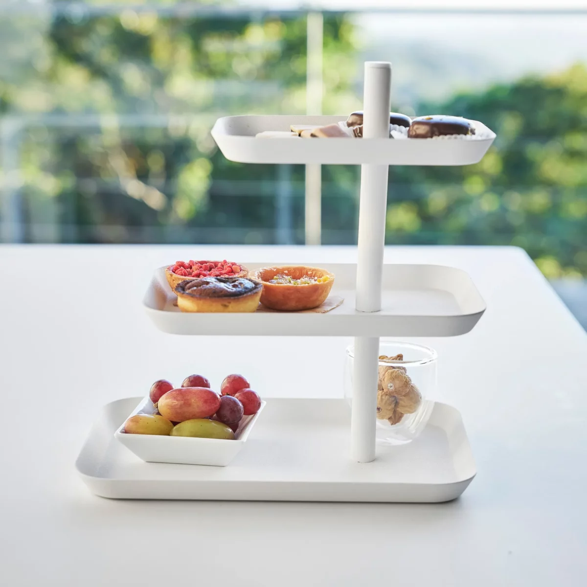 Yamazaki Etagere TOWER 3-stöckig drehbar in Weiss, von vorne vor dem Fenster mit Früchten und Törtchen