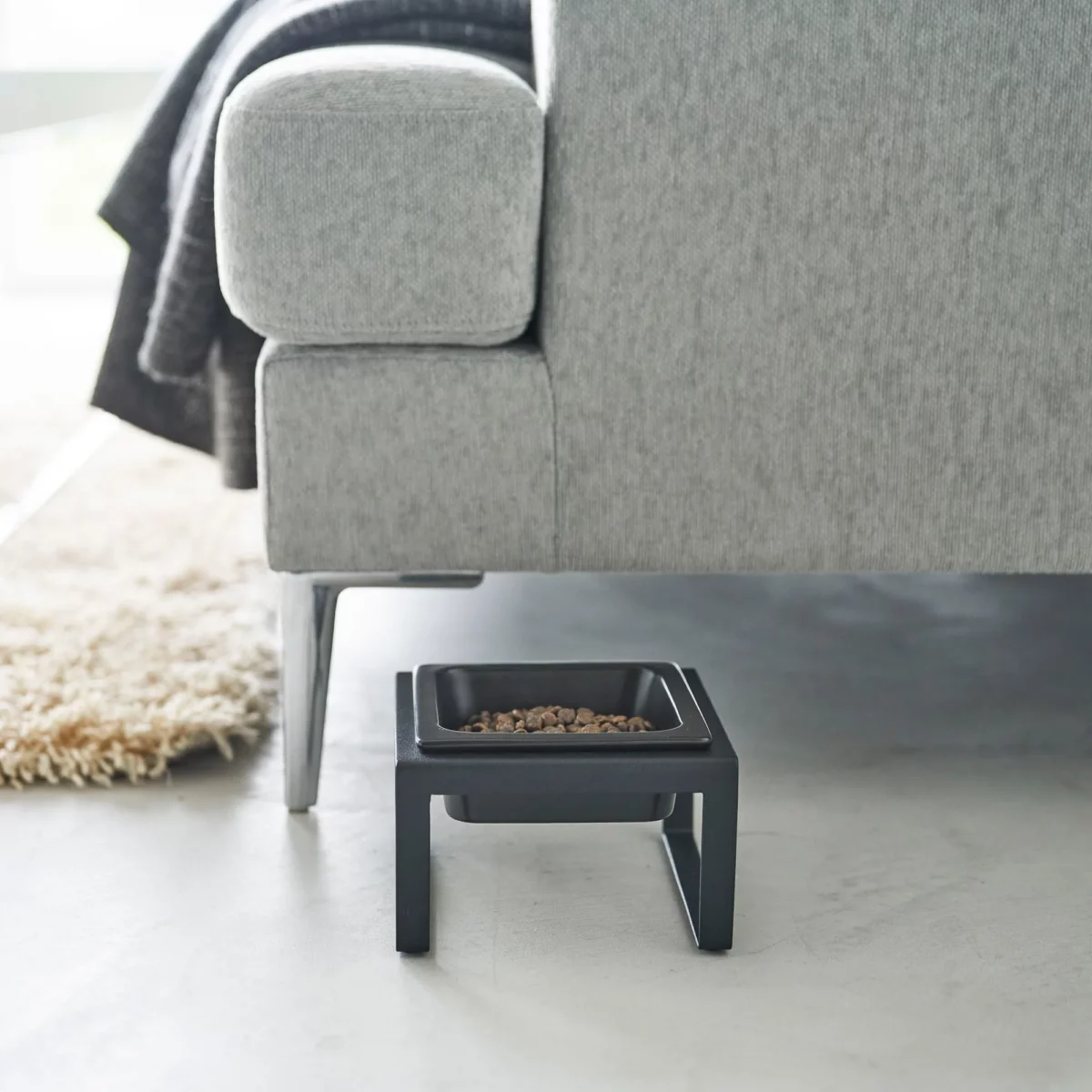 Yamazaki TOWER Food Bowl, single, with stand in black, on the concrete floor next to the sofa with food