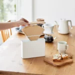 Yamazaki Kaffeefilterbox TOSCA mit Holzdeckel in Weiss, Hand nimmt einen Kaffeefilter aus der offenen Box