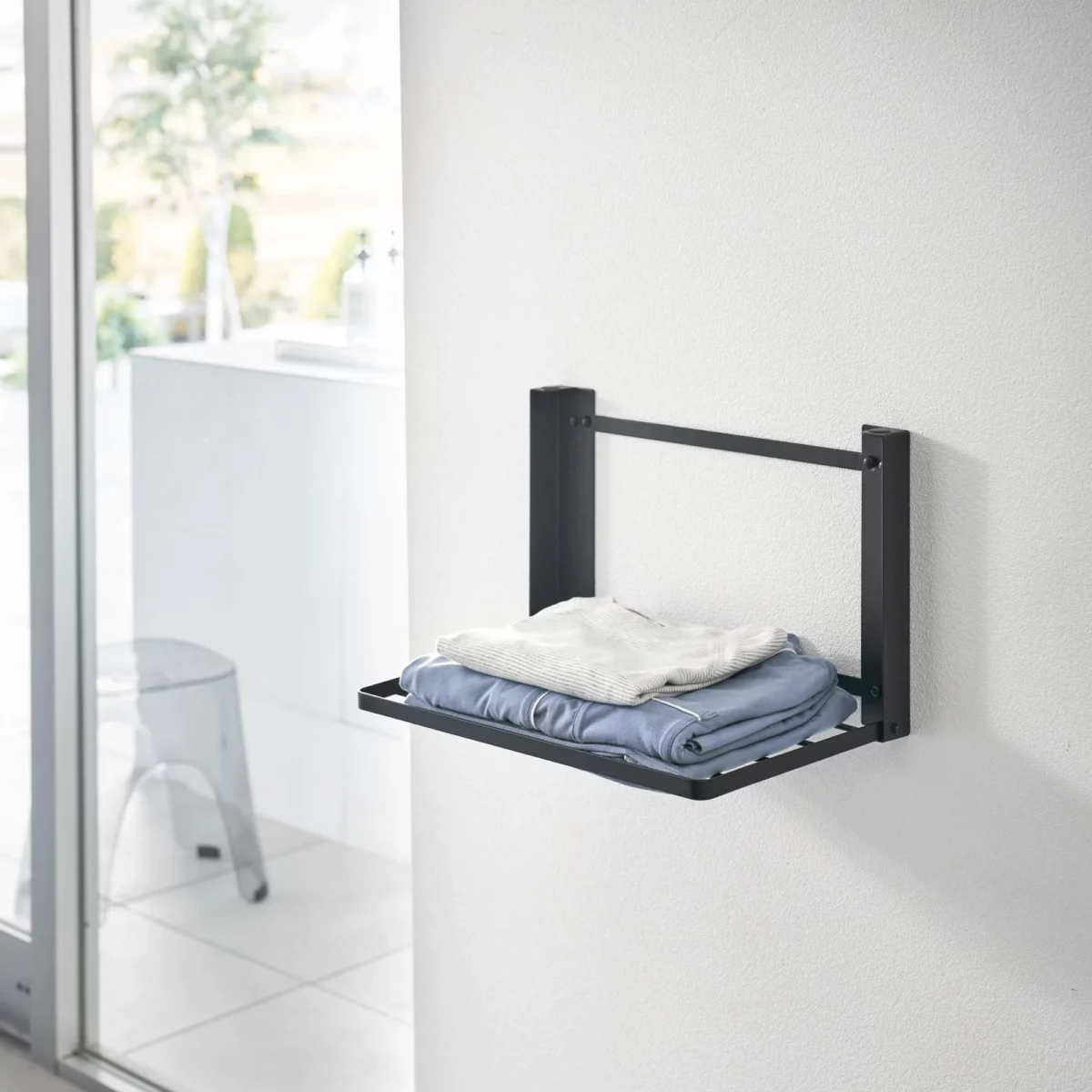 Yamazaki TOWER Folding Wall Shelf in Black, with folded clothes on the wall next to the window