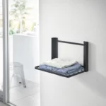 Yamazaki TOWER Folding Wall Shelf in Black, with folded clothes on the wall next to the window