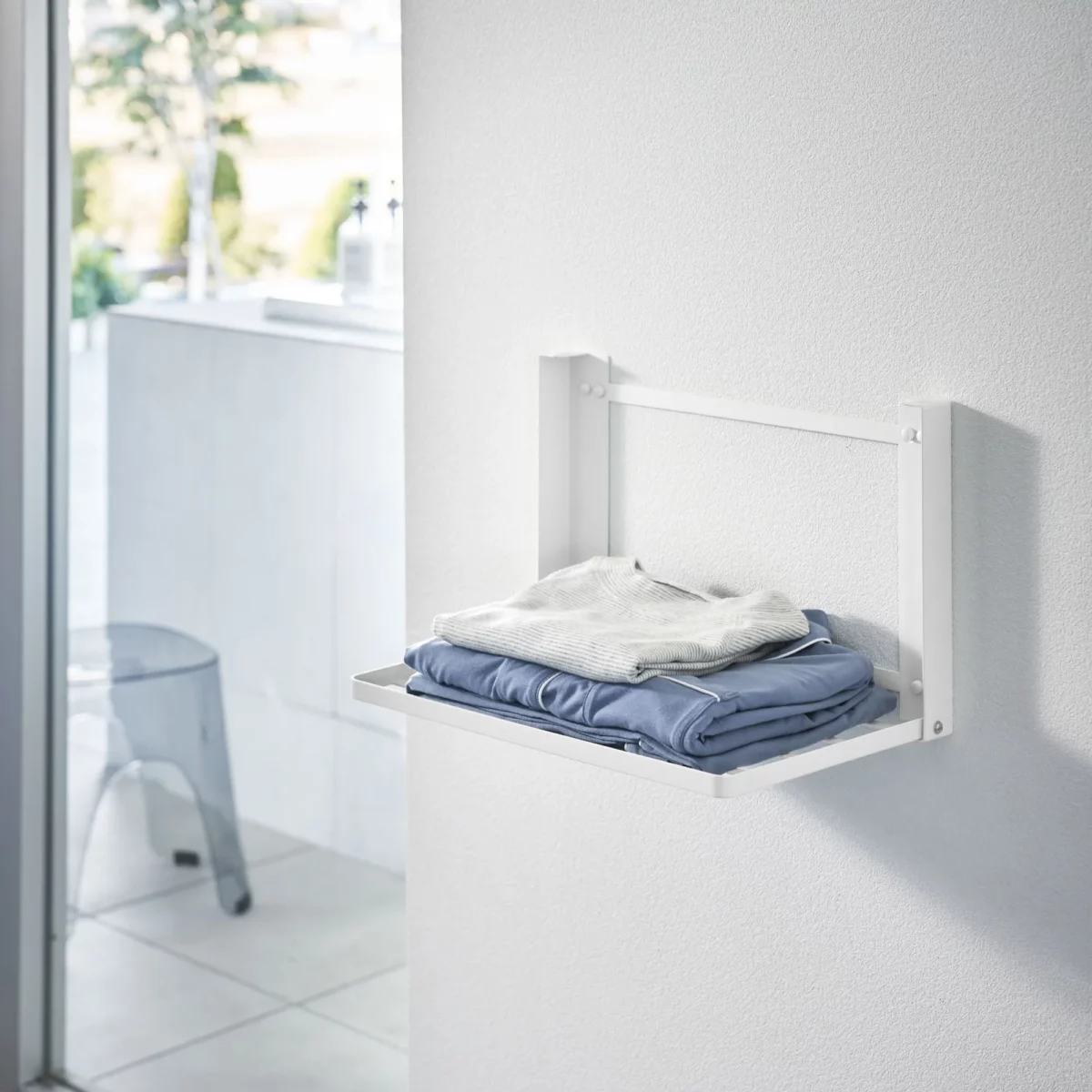 Yamazaki TOWER Folding Wall Shelf in White, with folded clothes on the wall next to the window