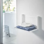 Yamazaki TOWER Folding Wall Shelf in White, with folded clothes on the wall next to the window