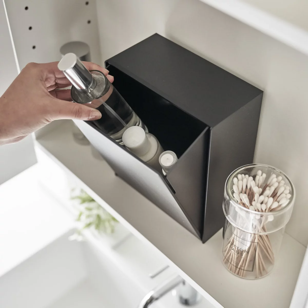 Boîte à crochets adhésifs Yamazaki TOWER avec couvercle, de couleur noire ; une main dépose une petite bouteille dans la boîte ouverte