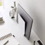 Support pour ordinateur portable Yamazaki TOWER vertical, blanc, avec un ordinateur portable posé sur le côté, à côté d'un réveil et de stylos