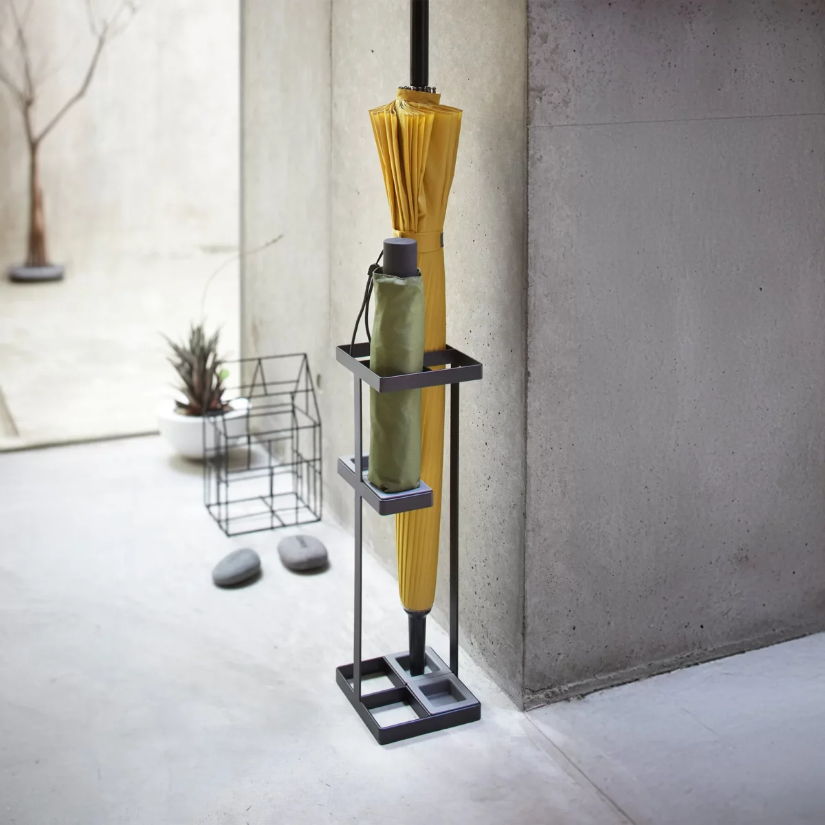 Yamazaki TOWER Slim Umbrella Stand in Black, against the concrete wall in the entryway with two umbrellas