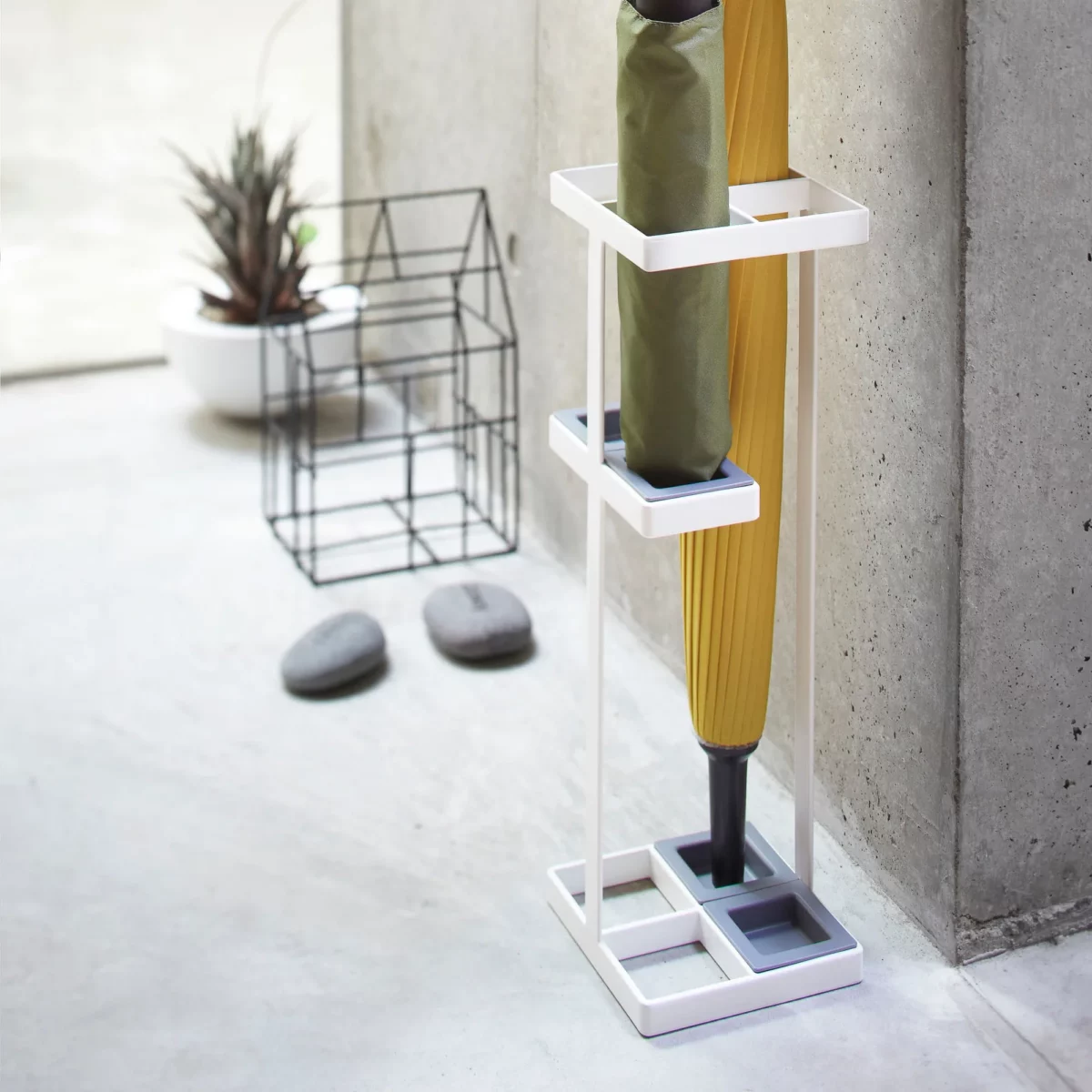 Yamazaki TOWER Slim Umbrella Stand in white, against the concrete wall in the entryway with two umbrellas