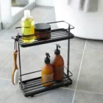 Étagère à distributeurs Yamazaki TOWER à 2 niveaux, de couleur noire, posée sur le sol de la salle de bains à côté de la baignoire, avec des produits de soin