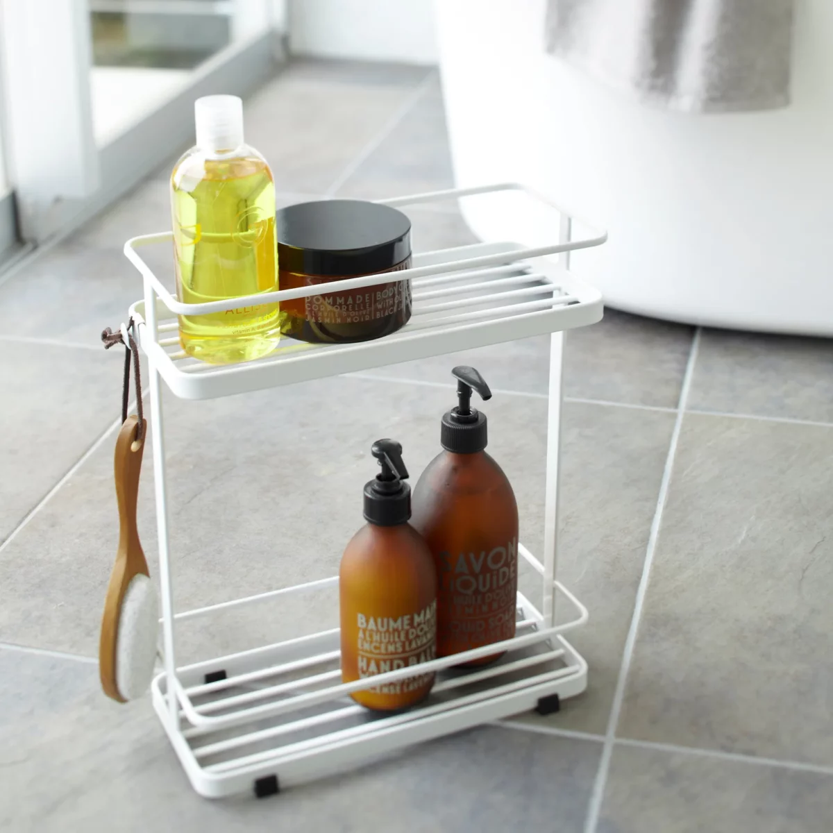 Étagère à distributeurs Yamazaki TOWER à 2 niveaux, de couleur blanche, posée sur le sol de la salle de bains à côté de la baignoire, avec des produits de soin