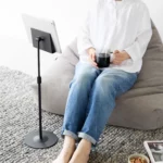 Support pour tablette Yamazaki SMART, réglable en hauteur, couleur noire ; une femme assise dans un pouf regarde sa tablette sans tenir l'appareil