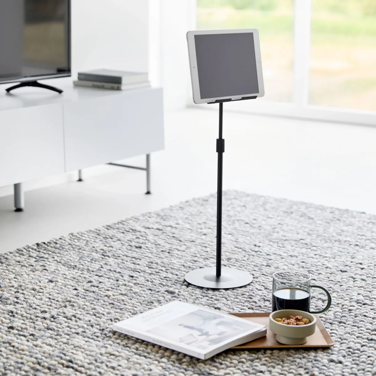 Support pour tablette Yamazaki SMART, réglable en hauteur, de couleur noire, posé au sol dans le salon, à côté d'un magazine et d'une tasse de thé
