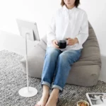Support pour tablette Yamazaki SMART réglable en hauteur, couleur blanche ; une femme assise dans un pouf regarde sa tablette sans tenir l'appareil