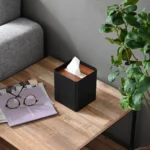 Yamazaki RIN square tissue box in black, on the wooden table next to glasses, a magazine, and a plant