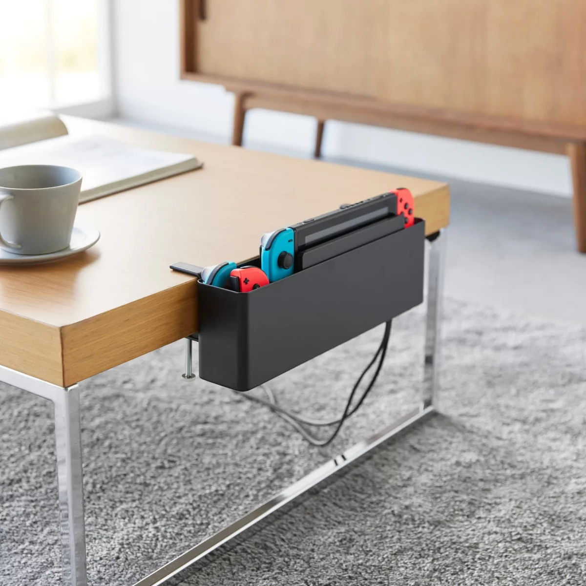 Yamazaki TOWER Narrow Table Clamp Basket in Black, on a coffee table with a game console and charging cable