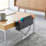 Yamazaki TOWER Narrow Table Clamp Basket in Black, on a coffee table with a game console and charging cable