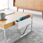 Yamazaki TOWER Narrow Table Clamp Basket in White, on a coffee table with a game console and charging cable