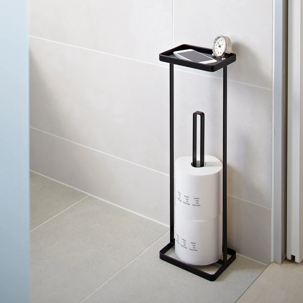 Yamazaki TOWER Toilet Paper Holder with Shelf in Black, mounted on the bathroom wall with toilet paper rolls, a clock, and a smartphone