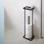 Yamazaki TOWER Toilet Paper Holder with Shelf in Black, mounted on the bathroom wall with toilet paper rolls, a clock, and a smartphone