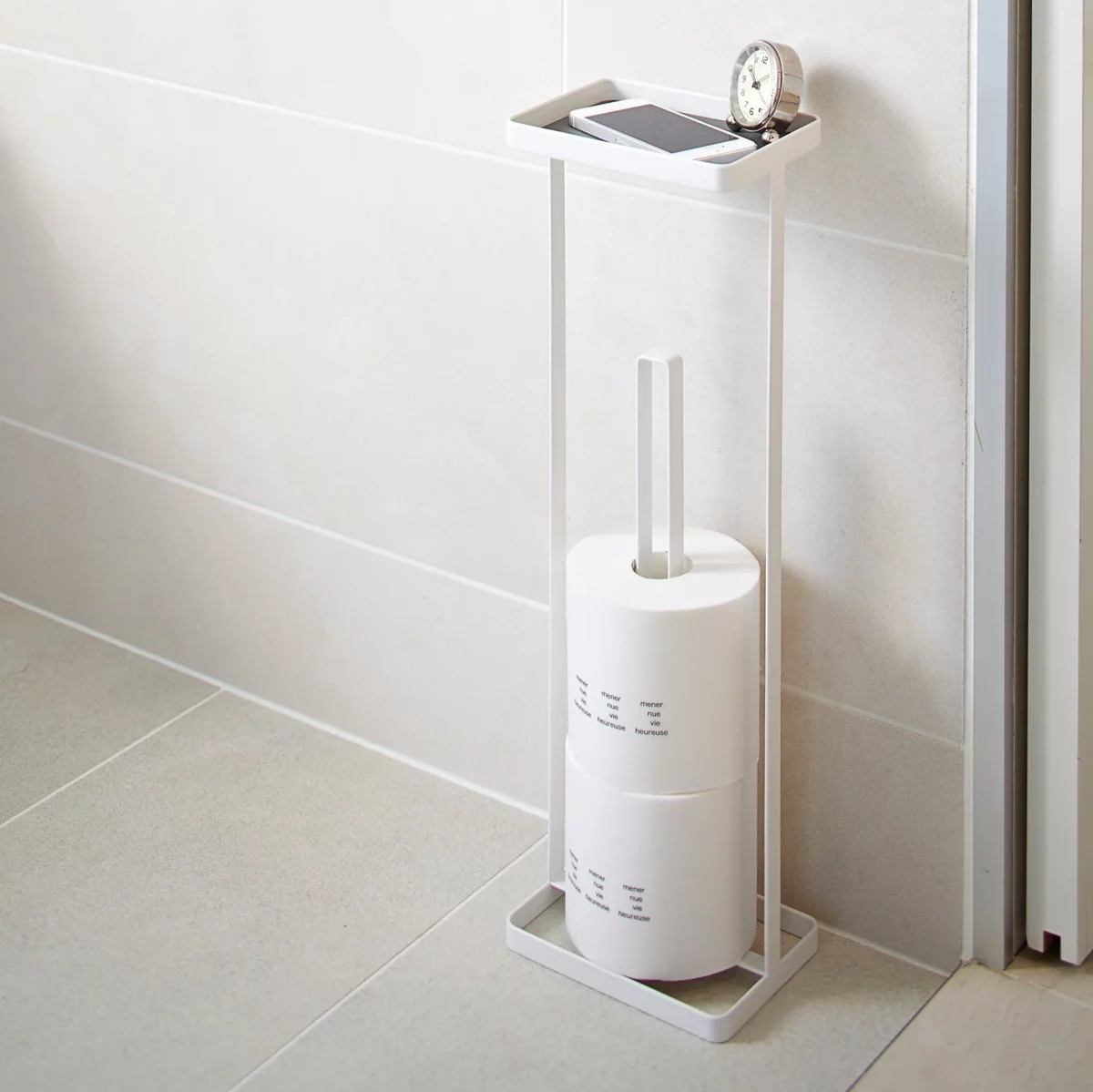 Yamazaki TOWER Toilet Paper Holder with Shelf in White, mounted on the bathroom wall with toilet paper rolls, a clock, and a smartphone