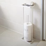 Yamazaki TOWER Toilet Paper Holder with Shelf in White, mounted on the bathroom wall with toilet paper rolls, a clock, and a smartphone