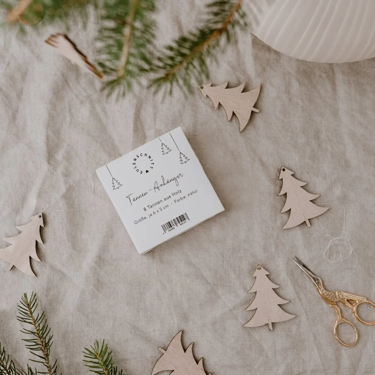 Eulenschnitt wooden fir tree ornament in natural finish, arranged on linen, with packaging, a fir branch, and gold scissors