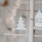 Eulenschnitt wooden ornament: a white fir tree on a wooden stick decorative tree next to a white house, with lights in the background